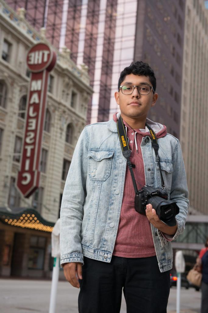Young man with camera in hand surrounded by tall buildings and a Majestic Theater sign in the background.