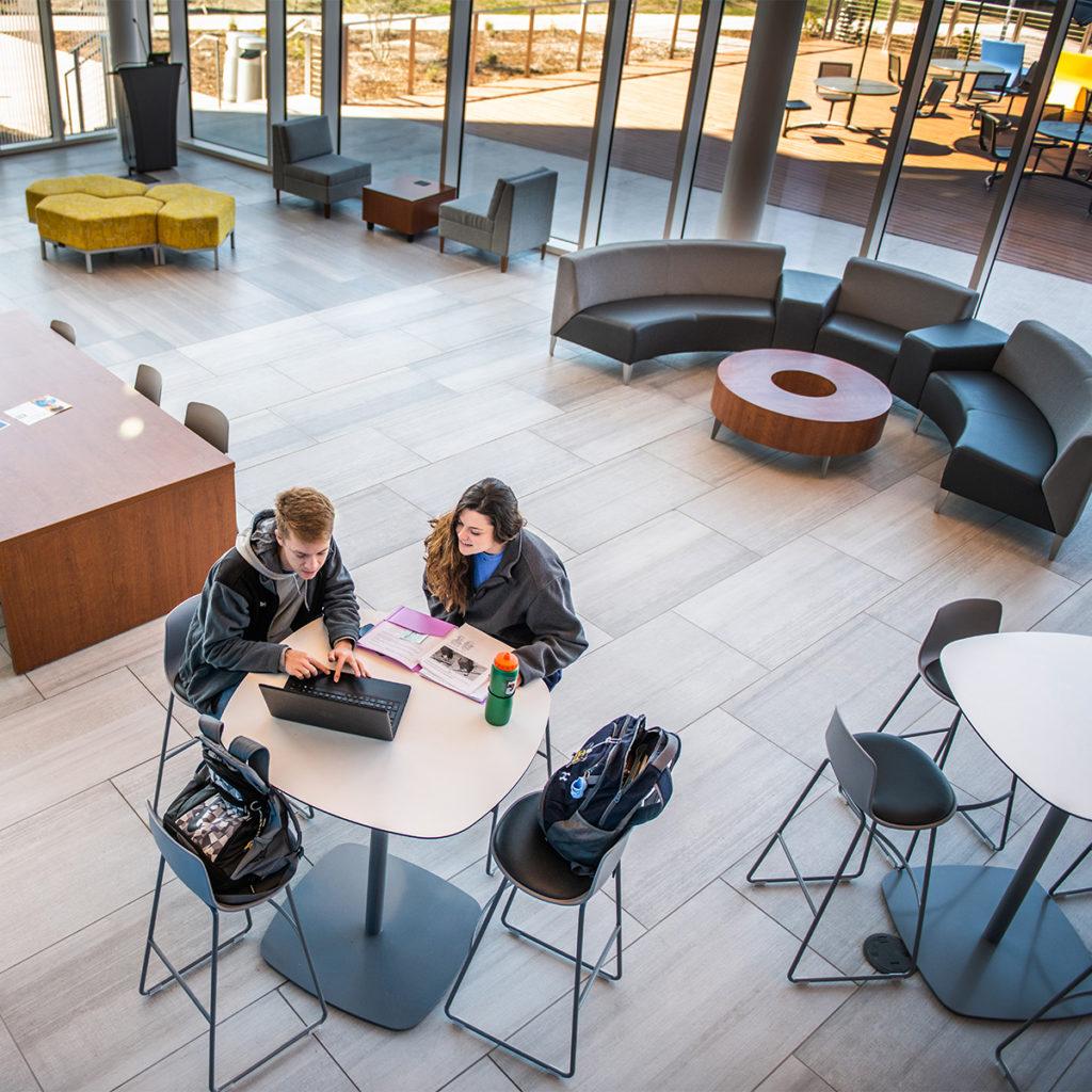 Two student sitting down at a table looking at a laptop in the common area of the nursing and health sciences building.