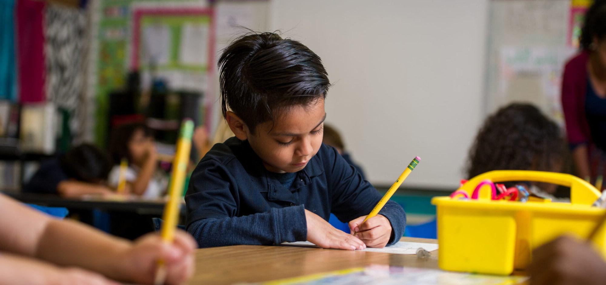 Young hispanic boy writing with pencil in classroom.